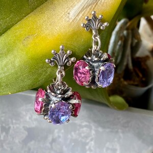 May include: A pair of silver dangle earrings with pink and purple gemstones. The earrings feature a floral design and a crown-shaped top.