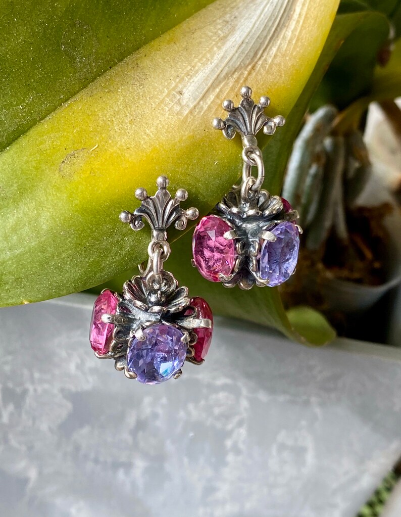May include: A pair of silver dangle earrings with pink and purple gemstones. The earrings feature a floral design and a crown-shaped top.