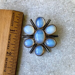 K&ouml;nnte beinhalten: Eine silberne Brosche mit einem Sternenausbruch-Design mit sieben schimmernden blauen ovalen Steinen.