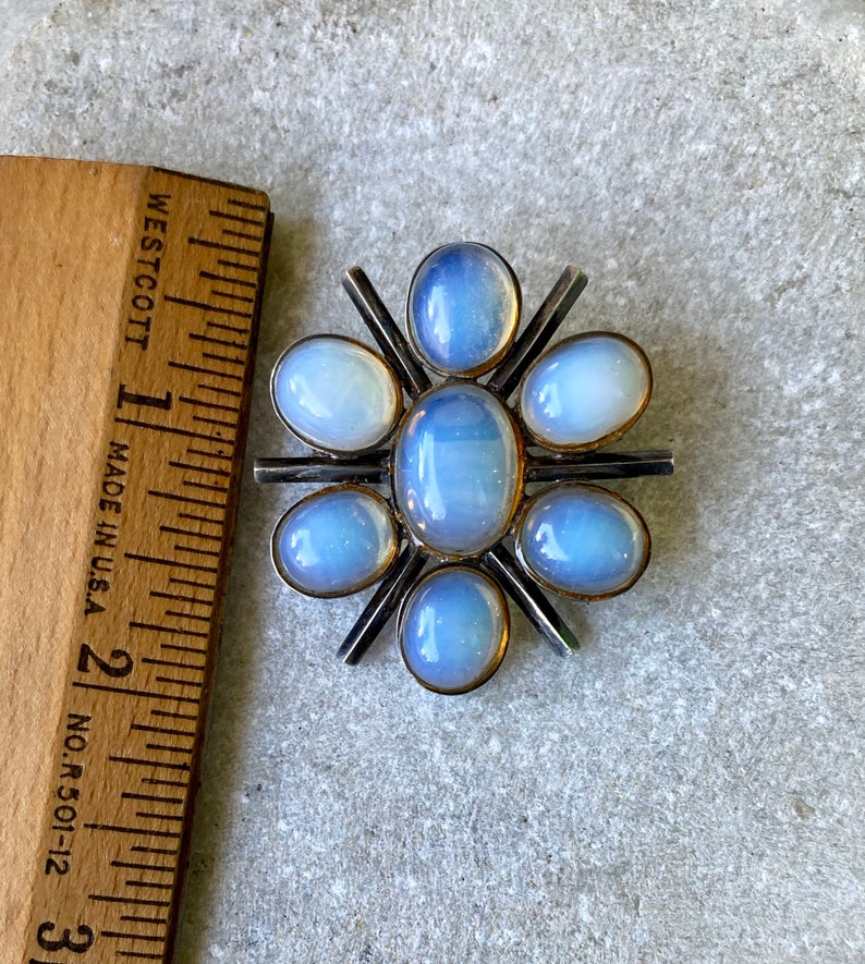 K&ouml;nnte beinhalten: Eine silberne Brosche mit einem Sternenausbruch-Design mit sieben schimmernden blauen ovalen Steinen.