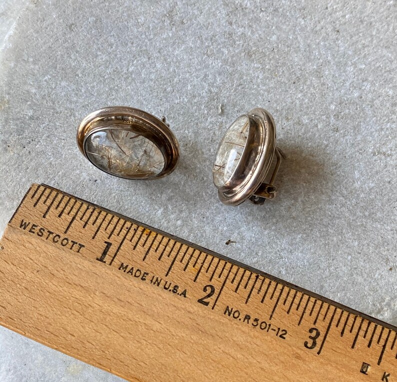 Puede incluir: Un par de pendientes de clip plateados vintage. Cada pendiente presenta un cabuj&oacute;n ovalado transparente con inclusiones doradas, engastado en un bisel plateado. Los pendientes miden aproximadamente 2 cm de largo.