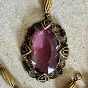May include: Antique-style pendant necklace featuring a large, oval, faceted purple gemstone. The gemstone is set in an ornate, bronze-toned metal frame with decorative accents and a matching chain.
