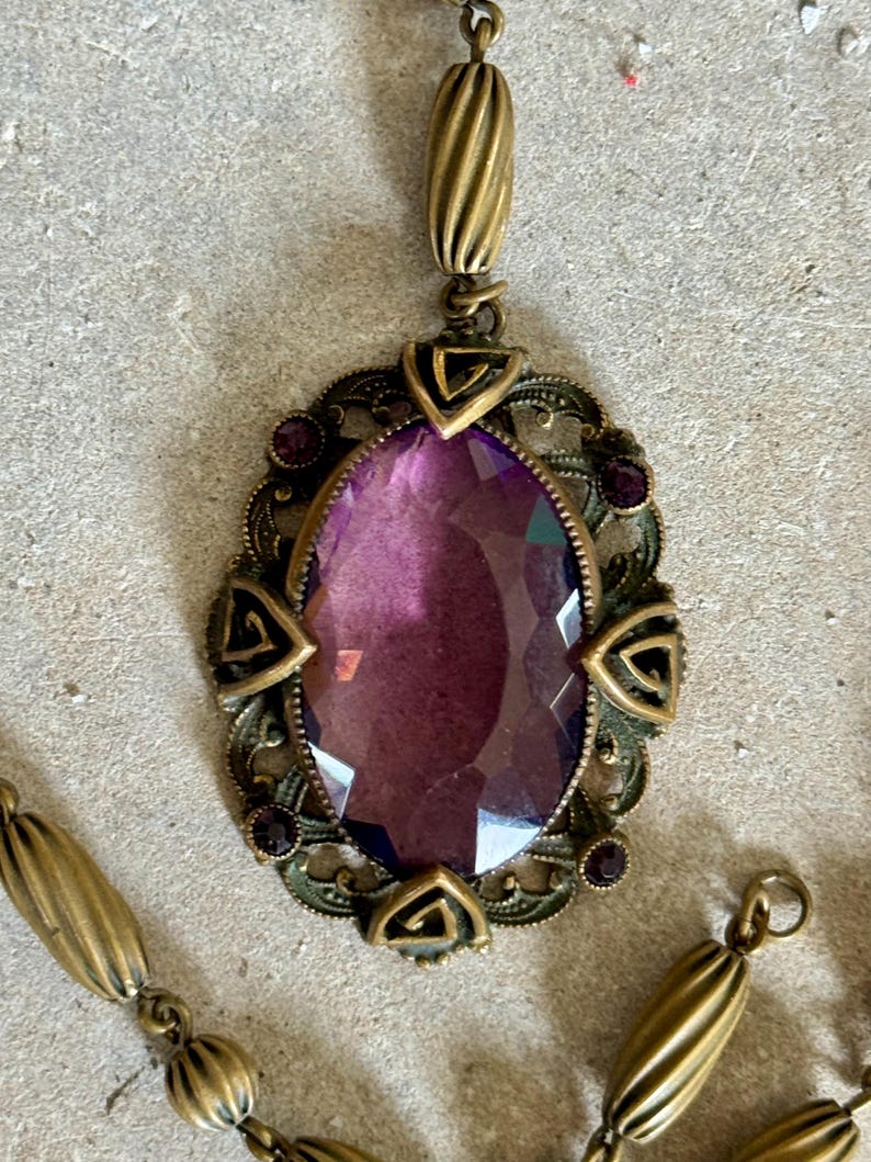 May include: Antique-style pendant necklace featuring a large, oval, faceted purple gemstone. The gemstone is set in an ornate, bronze-toned metal frame with decorative accents and a matching chain.