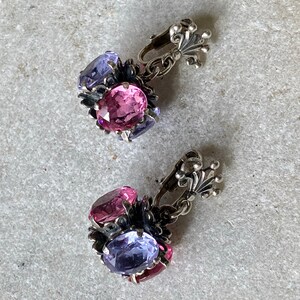 May include: A pair of silver clip-on earrings with pink and purple oval-shaped stones. The earrings have a floral design and a decorative silver clasp.