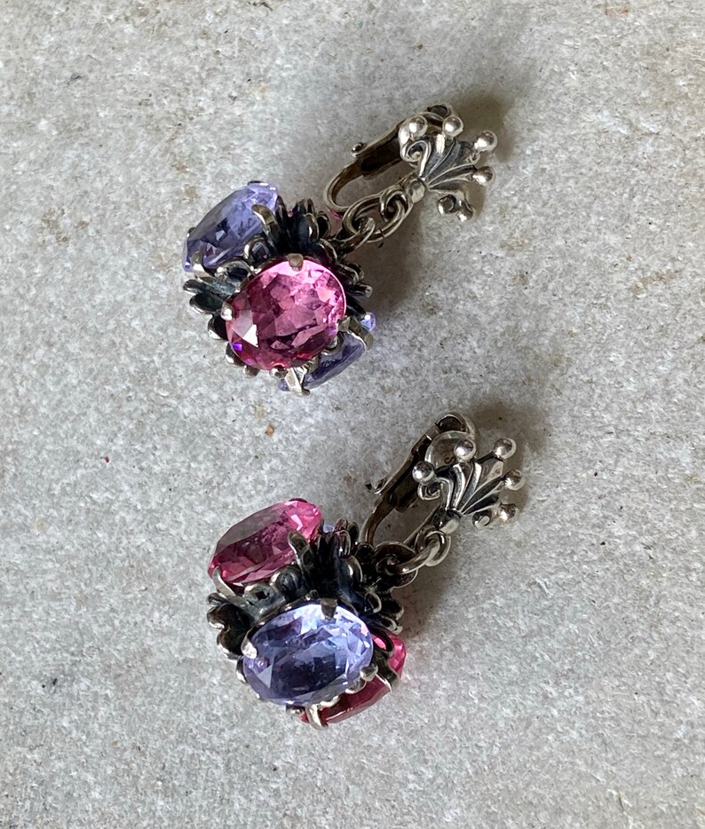 May include: A pair of silver clip-on earrings with pink and purple oval-shaped stones. The earrings have a floral design and a decorative silver clasp.