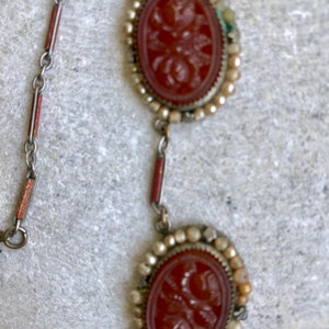 May include: A vintage necklace featuring two oval, reddish-brown cameo pendants with floral carvings, framed by small, off-white beads. The chain has silver links with red and silver accents. The necklace is displayed on a textured, light gray surface.