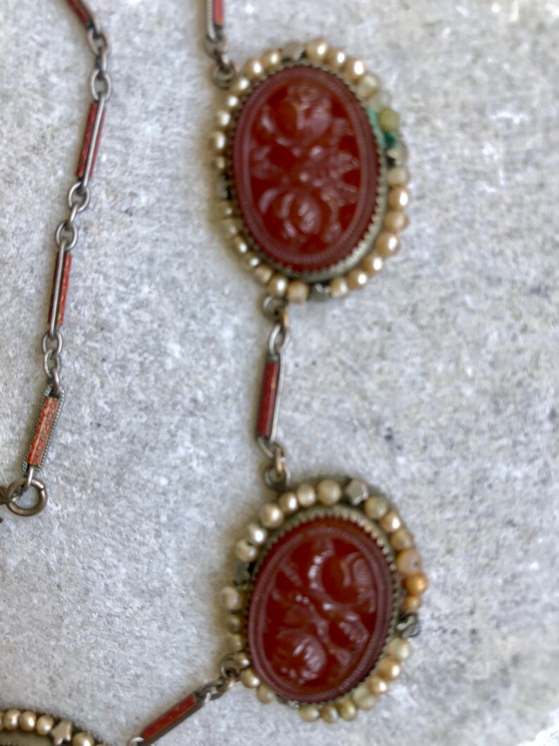 May include: A vintage necklace featuring two oval, reddish-brown cameo pendants with floral carvings, framed by small, off-white beads. The chain has silver links with red and silver accents. The necklace is displayed on a textured, light gray surface.