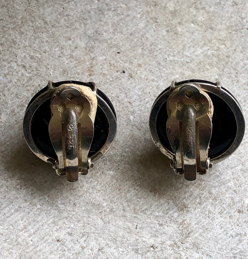 May include: A pair of black and silver clip-on earrings with a round, black stone. The earrings are marked with "925" on the back.