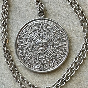 May include: A silver necklace with a round pendant featuring a detailed design of the Aztec calendar. The chain is a double chain with a delicate link.
