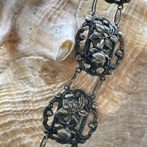 May include: A silver chain necklace with intricate floral and leaf designs. Each link features a detailed cutout of a flower and leaves.