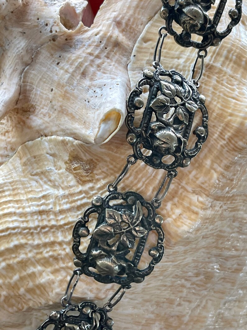 May include: A silver chain necklace with intricate floral and leaf designs. Each link features a detailed cutout of a flower and leaves.