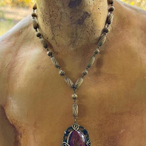 May include: Antique-style necklace with a bronze-toned chain and pendant. The pendant features a large, oval-shaped, deep purple gemstone set within an ornate, filigree frame. The necklace is displayed on a bust.