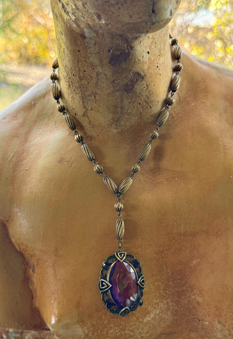May include: Antique-style necklace with a bronze-toned chain and pendant. The pendant features a large, oval-shaped, deep purple gemstone set within an ornate, filigree frame. The necklace is displayed on a bust.