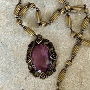 May include: Vintage brass-toned necklace featuring an oval purple gemstone pendant. The pendant is framed by an ornate, filigree-style setting with small purple accents. The necklace chain is composed of ribbed, barrel-shaped beads.