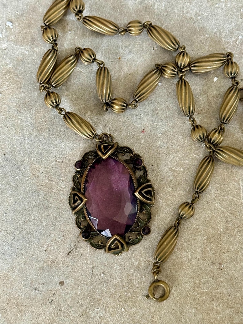 May include: Vintage brass-toned necklace featuring an oval purple gemstone pendant. The pendant is framed by an ornate, filigree-style setting with small purple accents. The necklace chain is composed of ribbed, barrel-shaped beads.