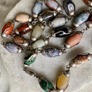 May include: A silver chain necklace with oval-shaped gemstone beads in various colors and patterns. The beads are set in silver and have a polished finish.
