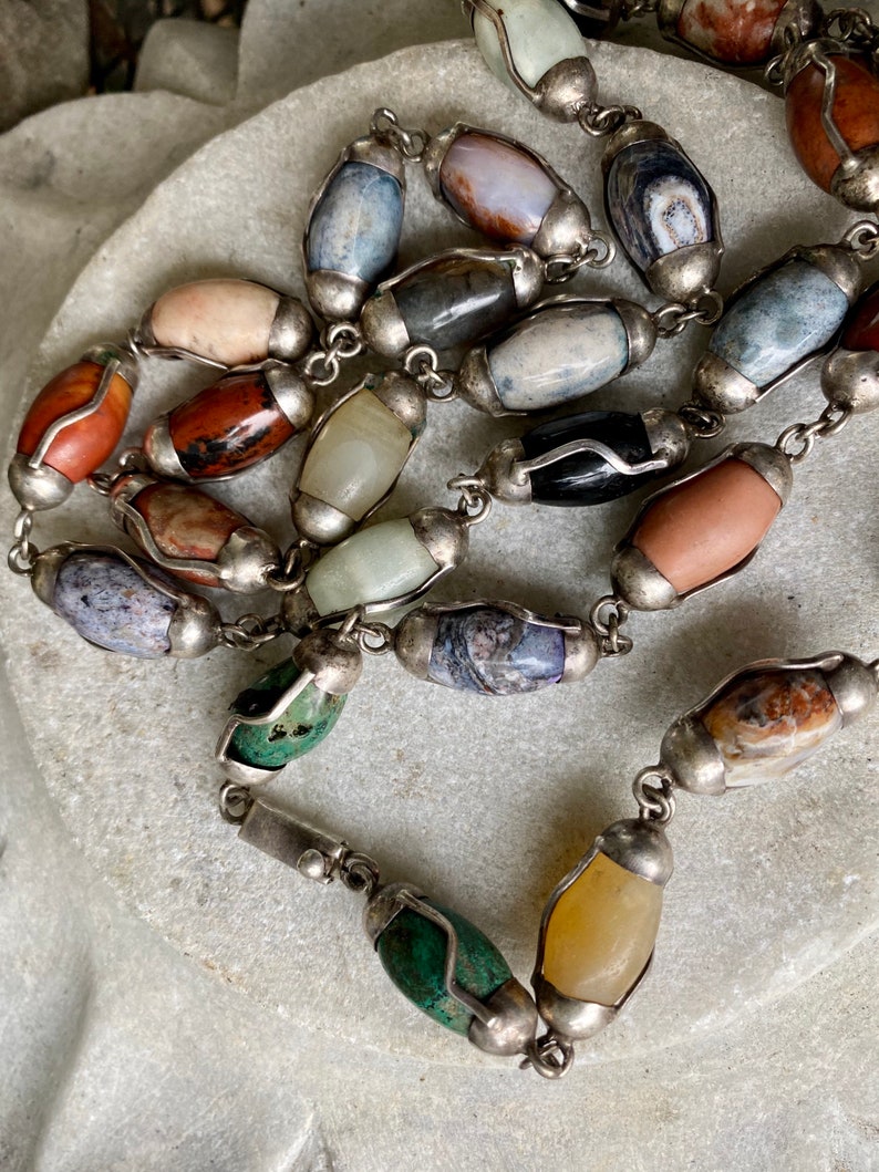 May include: A silver chain necklace with oval-shaped gemstone beads in various colors and patterns. The beads are set in silver and have a polished finish.