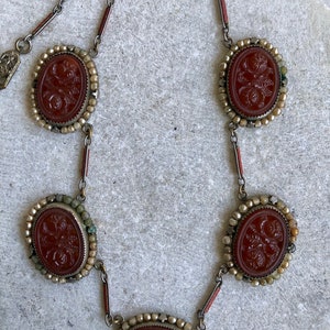 May include: A vintage necklace featuring five oval, reddish-brown carved stone pendants with floral designs. The pendants are set in silver-toned metal with a beaded border and connected by delicate chain links. The necklace has a classic, antique aesthetic.