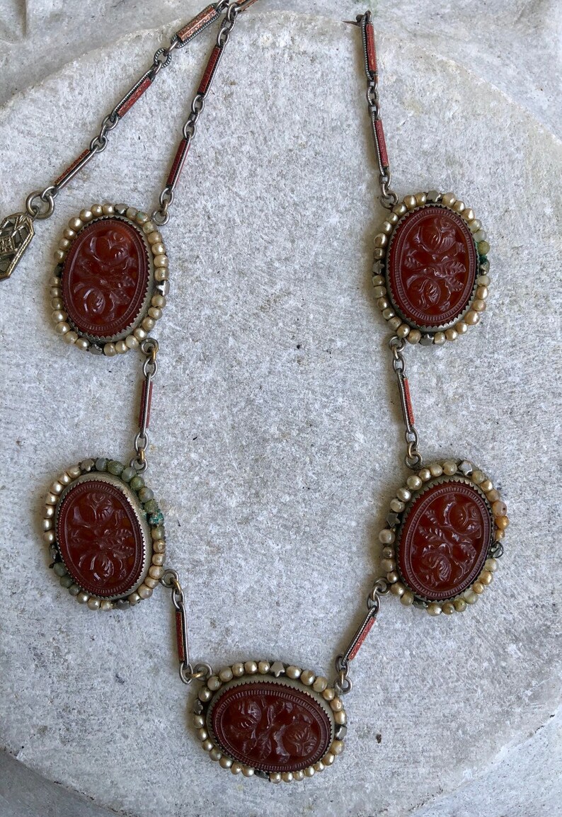 May include: A vintage necklace featuring five oval, reddish-brown carved stone pendants with floral designs. The pendants are set in silver-toned metal with a beaded border and connected by delicate chain links. The necklace has a classic, antique aesthetic.