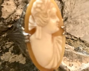 May include: Oval cameo ring featuring a profile of a woman in a cream-colored relief. The ring has a silver-colored metal band with decorative leaf-like accents on the sides. The cameo is set against a neutral background.