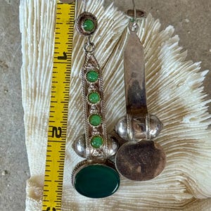 Pu&ograve; includere: Un paio di orecchini color argento con dettagli di pietre preziose verdi. Ogni orecchino presenta una grande pietra verde ovale alla base, con pietre verdi pi&ugrave; piccole ed elementi decorativi argentati. Gli orecchini sono lunghi circa 7,6 cm.