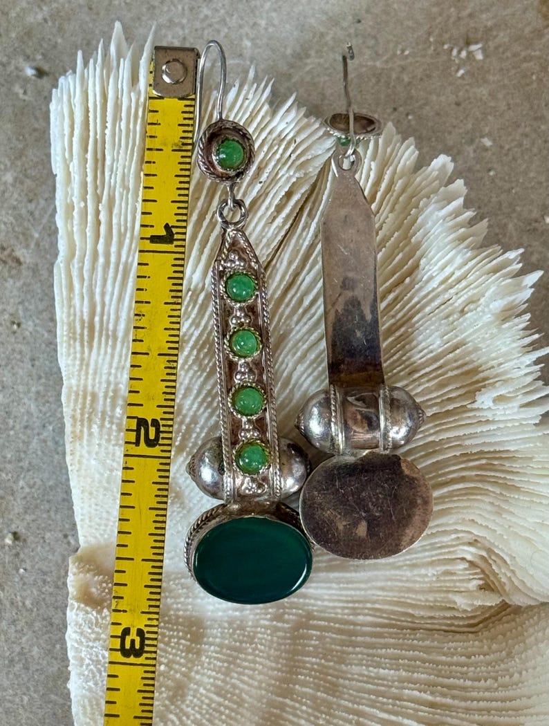 Pu&ograve; includere: Un paio di orecchini color argento con dettagli di pietre preziose verdi. Ogni orecchino presenta una grande pietra verde ovale alla base, con pietre verdi pi&ugrave; piccole ed elementi decorativi argentati. Gli orecchini sono lunghi circa 7,6 cm.