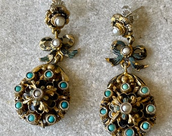 May include: A pair of gold tone earrings with turquoise and pearl accents. The earrings feature a floral design with a bow at the top.