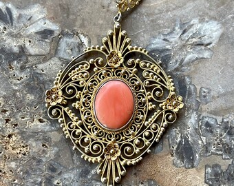 May include: A delicate gold filigree pendant with a pink coral stone set in the centre. The pendant features intricate floral designs and a dangling drop.