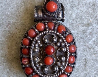 May include: An oval-shaped pendant with a silver filigree design and a border of red coral beads. The pendant has a loop at the top for hanging.
