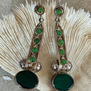 Pu&ograve; includere: Un paio di orecchini pendenti argentati. Ogni orecchino presenta una grande pietra ovale verde scuro nella parte inferiore, con pietre verdi pi&ugrave; piccole ed elementi decorativi argentati. Gli orecchini sono esposti su uno sfondo strutturato bianco sporco.