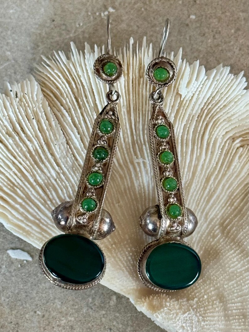 Pu&ograve; includere: Un paio di orecchini pendenti argentati. Ogni orecchino presenta una grande pietra ovale verde scuro nella parte inferiore, con pietre verdi pi&ugrave; piccole ed elementi decorativi argentati. Gli orecchini sono esposti su uno sfondo strutturato bianco sporco.