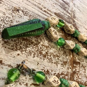 以下が含まれることがあります： 緑と白のビーズネックレスで、緑色のスカラベのペンダントが付いています。ビーズは骨とガラスでできています。ネックレスは留め金で留めることができます。