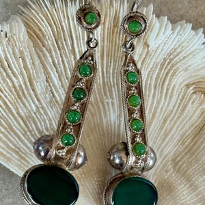 Pu&ograve; includere: Un paio di orecchini pendenti argentati. Ogni orecchino presenta una grande pietra ovale verde scuro nella parte inferiore, con una serie di pietre verdi pi&ugrave; piccole che salgono su una colonna rettangolare. La parte superiore ha una piccola pietra verde circondata da un motivo circolare.