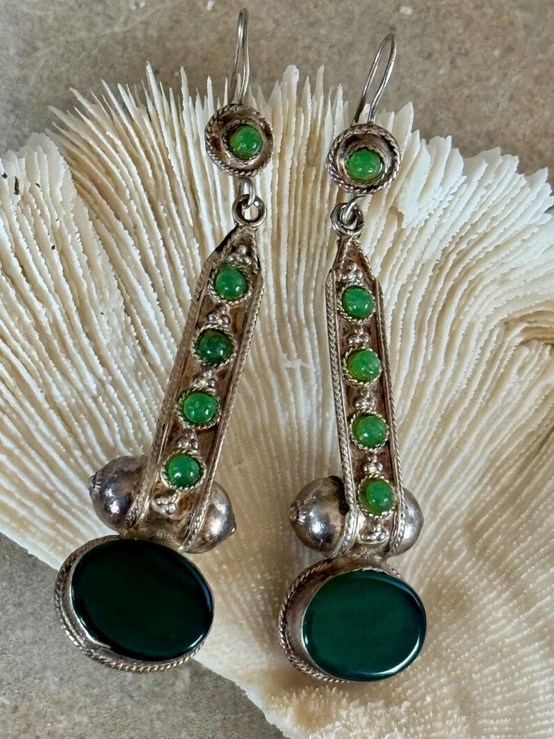 Pu&ograve; includere: Un paio di orecchini pendenti argentati. Ogni orecchino presenta una grande pietra ovale verde scuro nella parte inferiore, con una serie di pietre verdi pi&ugrave; piccole che salgono su una colonna rettangolare. La parte superiore ha una piccola pietra verde circondata da un motivo circolare.