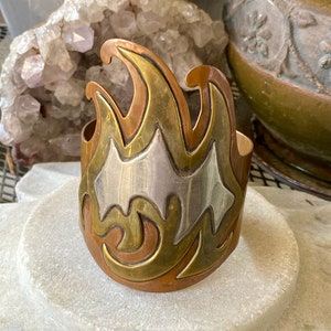 May include: A wide copper bracelet with a silver and brass design. The design is a stylized flame with a geometric pattern.