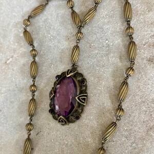 May include: Antique brass-toned necklace featuring an oval pendant with a large, faceted purple gemstone. The pendant is framed by an ornate, filigree-style setting with smaller purple accents. The chain is composed of alternating ribbed and round beads.