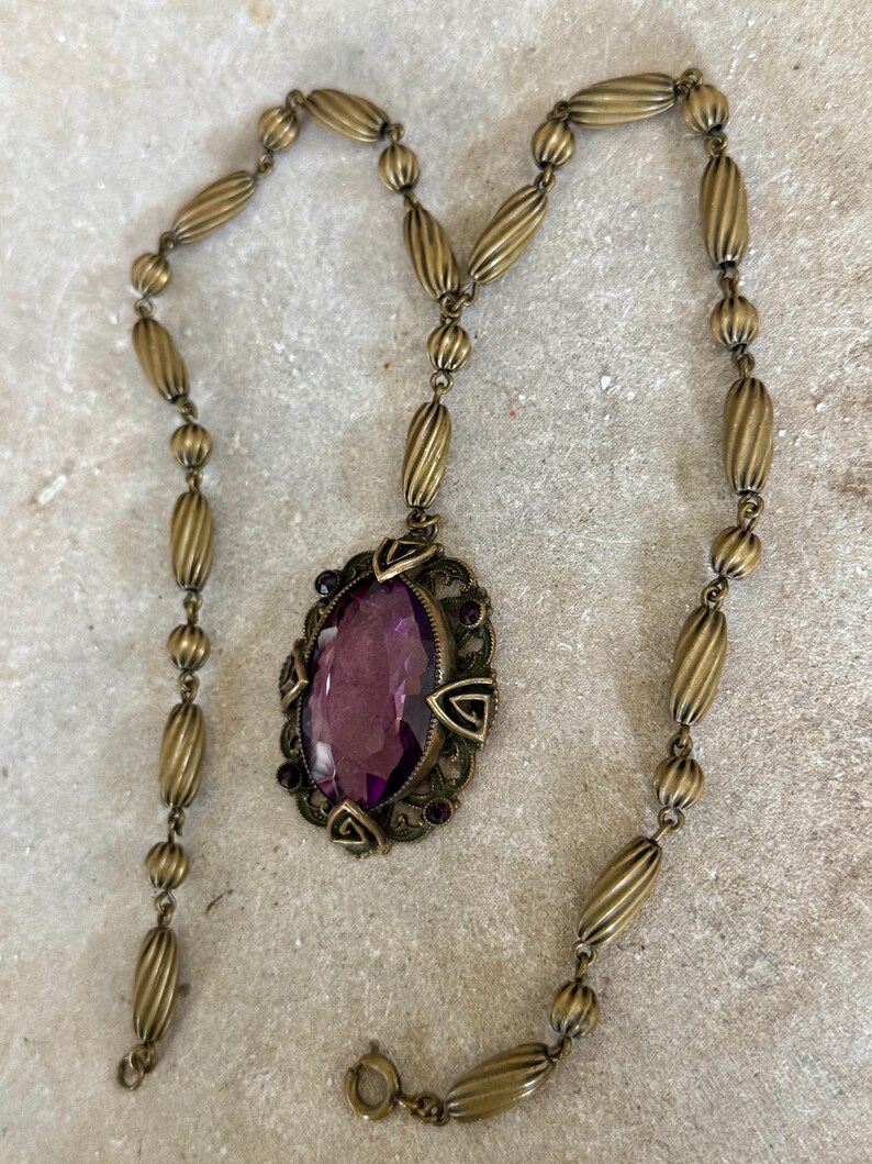May include: Antique brass-toned necklace featuring an oval pendant with a large, faceted purple gemstone. The pendant is framed by an ornate, filigree-style setting with smaller purple accents. The chain is composed of alternating ribbed and round beads.