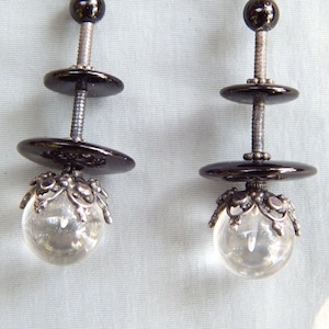 May include: A pair of earrings with black and silver accents. Each earring features two black round discs and a clear crystal ball with a silver filigree design.