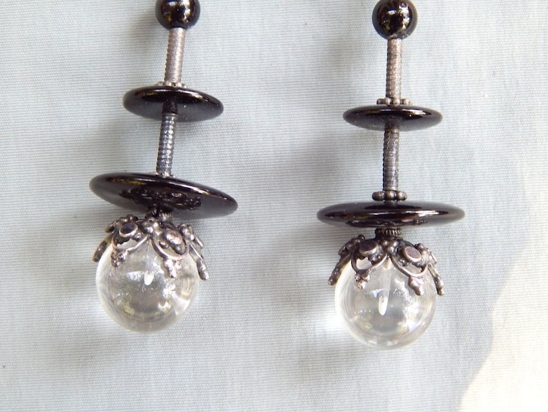 May include: A pair of earrings with black and silver accents. Each earring features two black round discs and a clear crystal ball with a silver filigree design.