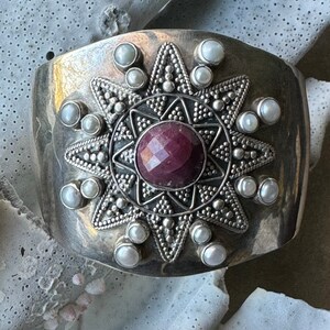May include: A silver cuff bracelet with a large, faceted ruby gemstone surrounded by a starburst design and white pearls.