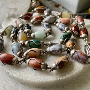 May include: A silver chain necklace with oval-shaped gemstone beads in various colors, including gray, brown, green, and white.
