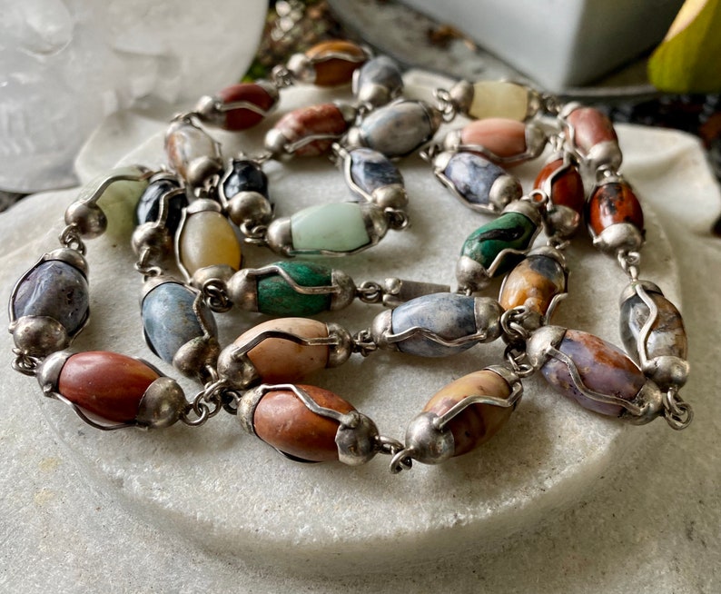 May include: A silver chain necklace with oval-shaped gemstone beads in various colors, including gray, brown, green, and white.