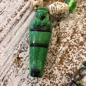 以下が含まれることがあります： 緑色のガラス製のペンダントは、中に彫刻された人物像が入ったエジプトの石棺の形をしています。ペンダントは、白と緑のビーズのネックレスの一部です。