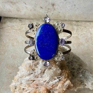 May include: A silver bracelet with a large oval lapis lazuli stone surrounded by smaller amethyst stones. The bracelet has a decorative design with swirls and is set on a bed of clear quartz crystals.