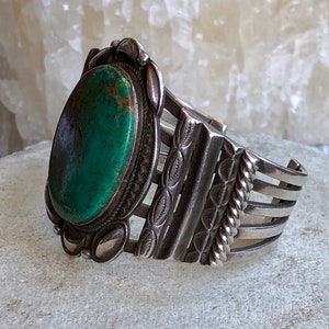 Puede incluir: Un brazalete de plata con una gran piedra turquesa ovalada engastada en el centro. El brazalete tiene un dise&ntilde;o decorativo con m&uacute;ltiples bandas y un patr&oacute;n de cuerda.