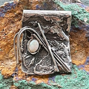 May include: A silver pendant with a textured surface and a white opal stone. The pendant is set against a background of brown and green rock.