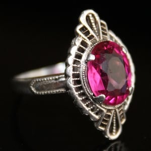 May include: A silver ring with a large, oval-shaped, bright pink gemstone. The gemstone is set in a decorative, filigree-style setting with detailed patterns. The ring's band is silver and has a simple, elegant design.