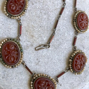 May include: A vintage necklace featuring oval, reddish-brown cameo pendants with floral carvings. The cameos are set in ornate frames with small, round beads and connected by a silver-toned chain with reddish-brown accents. A clasp secures the necklace.