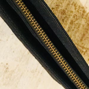 May include: Close-up of a black zipper with gold teeth. The zipper is sewn into a black fabric pouch.