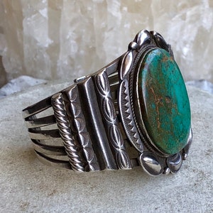 Puede incluir: Un brazalete de plata con una gran piedra ovalada de turquesa engastada en un engaste de plata. El brazalete tiene un dise&ntilde;o decorativo con m&uacute;ltiples bandas y una bisagra.